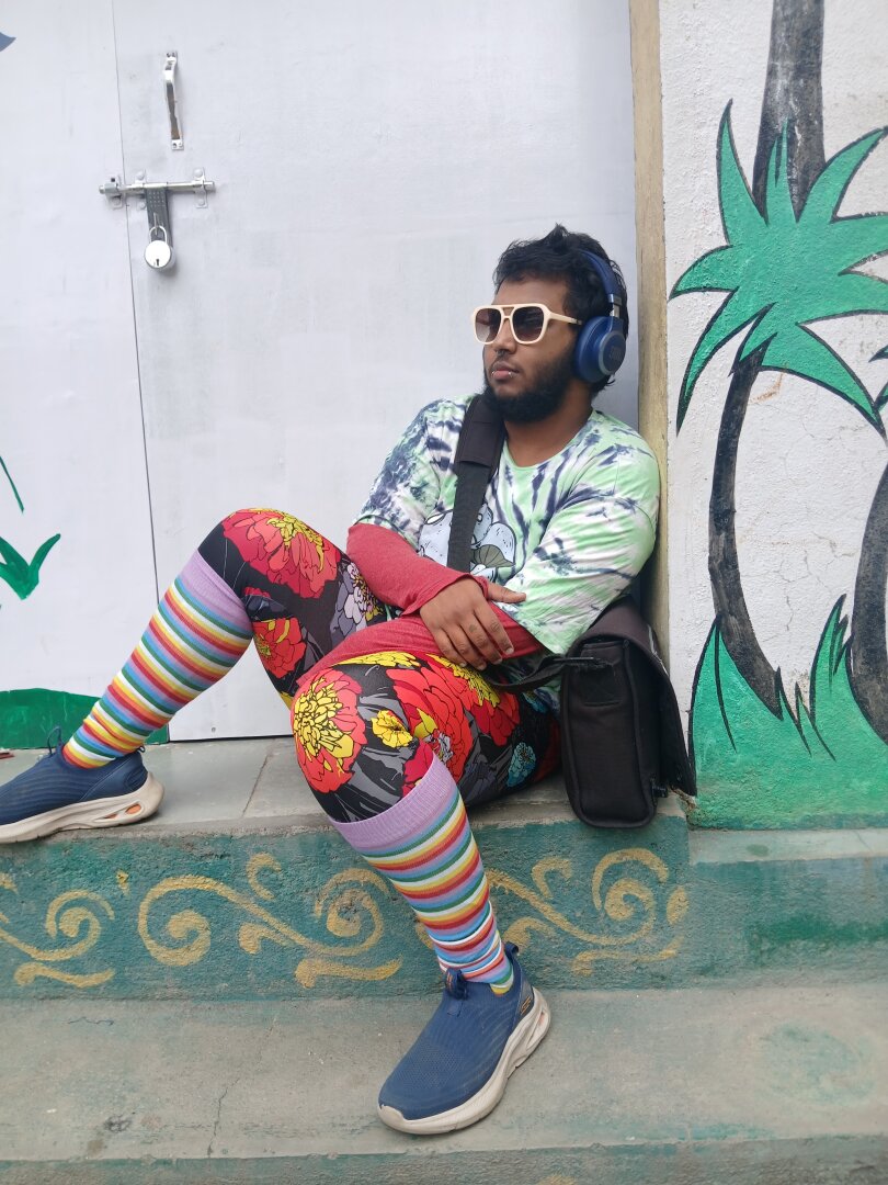 Image of a dark skinned brown person with black hair -  a thick beard and a thin moustache, sitting by a painted wall, leaning on a white locked door. they are wearing sunglasses with pastel pink frames, have blue headphones on, and have a satchel they're holding. they are wearing: blue slip on sneakers, athletic cycling pants with red and yellow flowers on them, knee high rainbow socks, a red full sleeved tshirt over which they're wearing a pastel green and dark blue tie and die half sleeved tshirt which has pastel blue mushrooms on it. their expression is neutral and they are gazing into the distance.