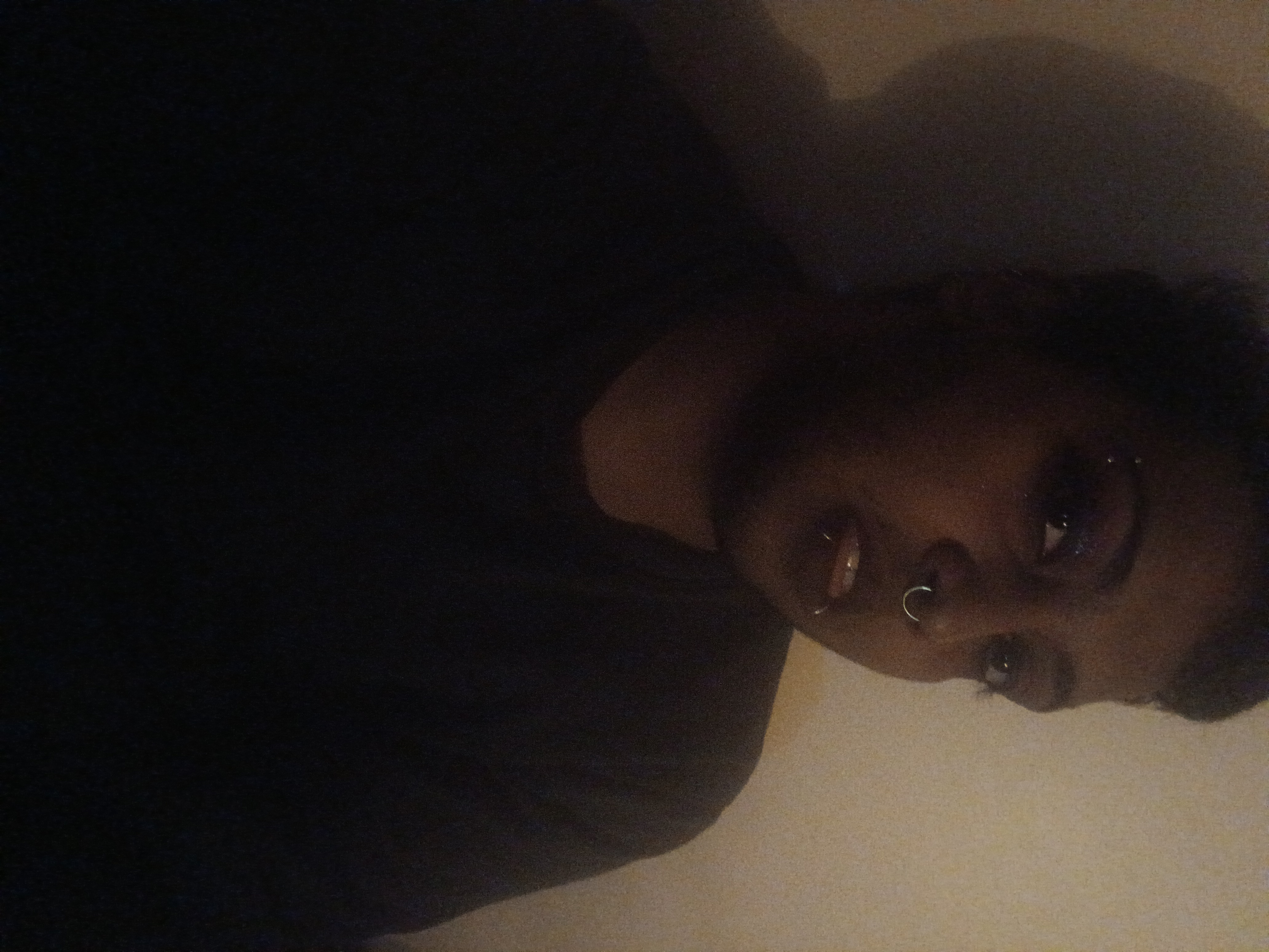 image of me, a dark skinned brown man's face and bust. i am in shadow in a dimly lit room but my makeup (eyeliner, glittery eyeshadow, dark plum lipstick) are visible. the shadows makes it look gothic, and my piercings (one eyebrow piercing, my two snakebites, and my septum ring) all gleam in the dark.