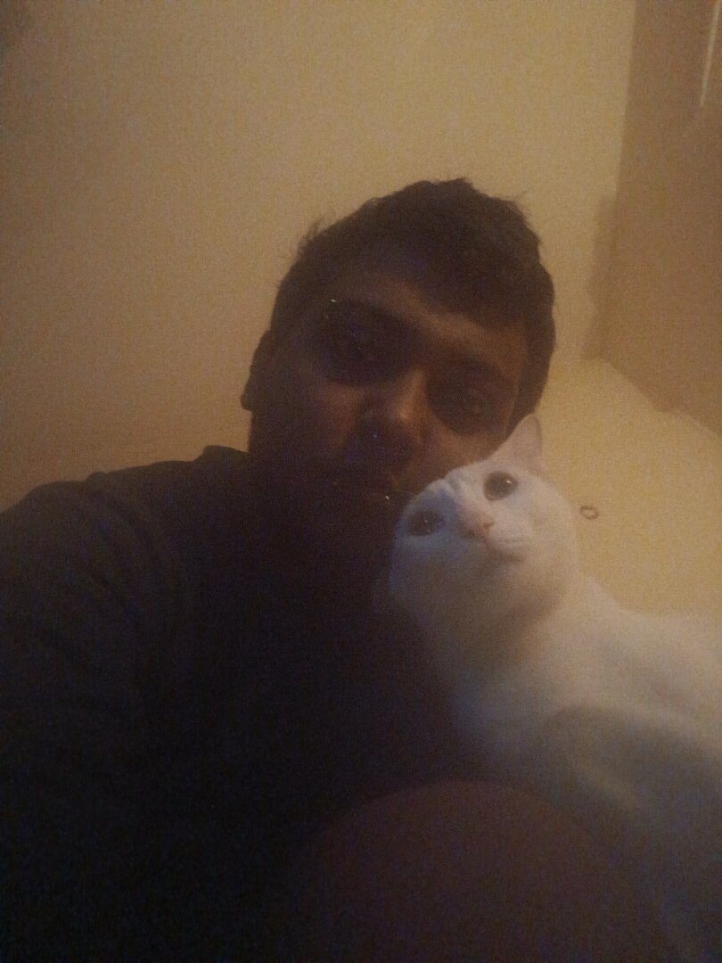 image of me (dark skinned brown man in a black turtleneck with gothic makeup) and my cat (fully white coat, green eyes and pink nose) staring in the direction of the camera - my expression is deadpan, whereas my cat looks like he is making pleading eyes like the emoji. our faces are pressed against each other, and one of his ears looks flappy because of the angle.