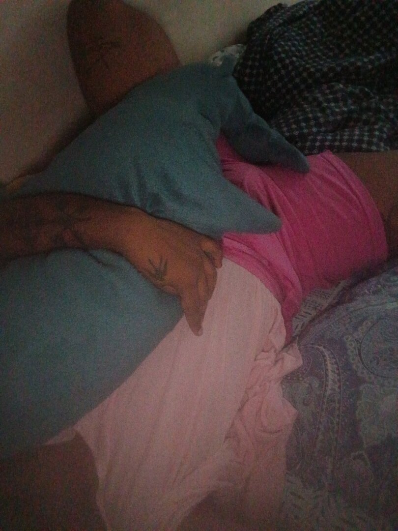 image of the torso and thighs of a dark skinned brown person wearing a pastel pink tshirt and hot pink pants, one of their arms are holding a blahaj close to their body. other than the hand holding the blahaj, no other hands/legs etc are visible, nor is their head visible