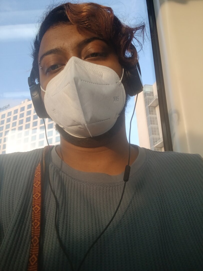 image of me, a dark skinned brown man, with a white n95 mask and headphones on, wearing a textured grey-blue tshirt. you can see skyscrapers and a blue sky through a glass pane behind me. a single red strand of hair is curling by the side of my face.