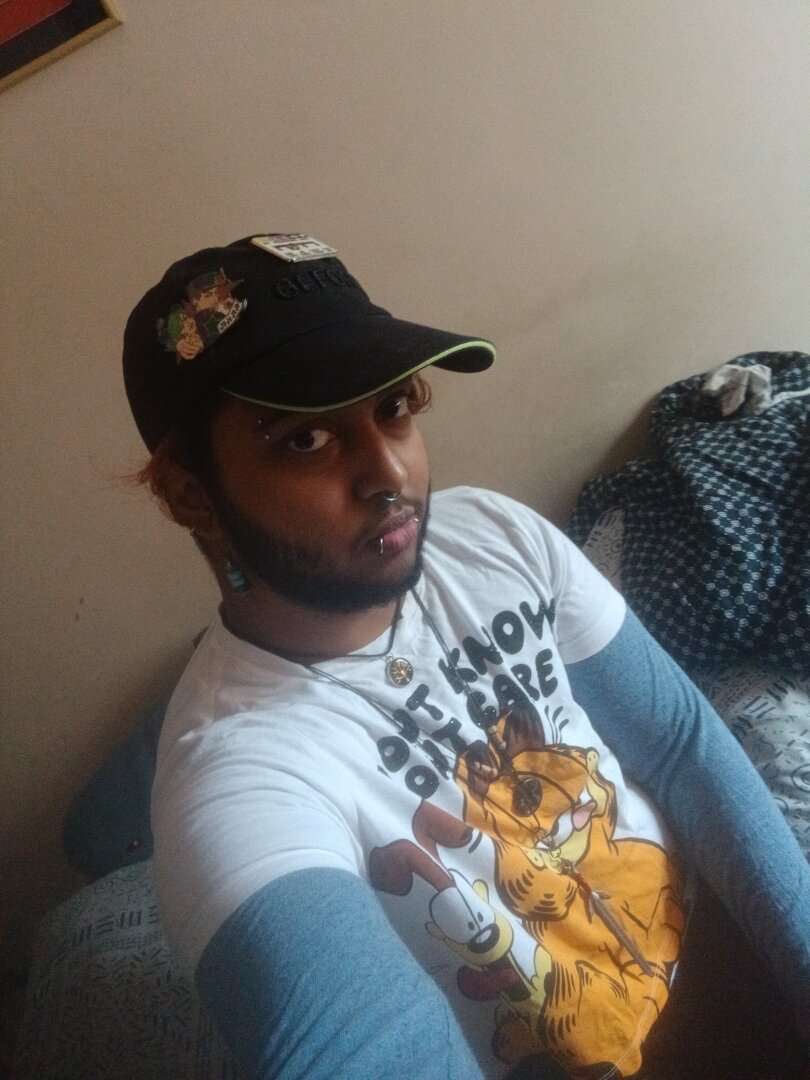 selfie of a dark skinned man wearing a white garfield t-shirt that has garfield and odie on it with the caption "don't know, don't care", with blue sleeves of a tshirt worn under it as a second layer. he is wearing a cap that has a jjba stone ocean pin on it. he is wearing a lot of jewelry - blue bead earrings, eyebrow piercing, septum piercing, snakebites, and three different necklaces featuring the wisdom tree, moon-face, sword from the witcher, and he has a beard and slight moustache. he is staring at the camera.
