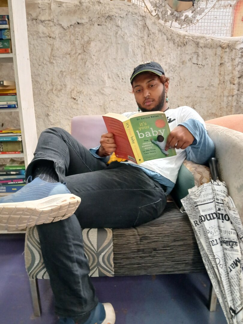 Image of me, a dark skinned brown man, sitting on a couch with multipatterned upholestry, and reading a book called "it's your baby" by Dr. Saroja Balan. the book is multicolored, and my posture and the camera angle make the image look silly despite my serious expression.