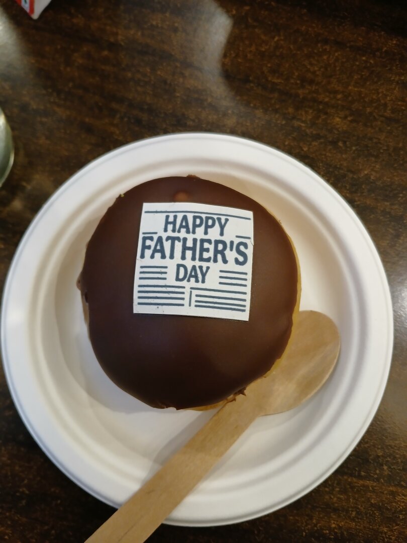 a chocolate donut on a paper plate with a wooden spoon. the donut has a fondant square on top of it that says "happy father's day" in capital letters.